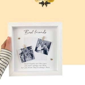 Hand holding a square white shadowbox photo frame that reads "Best friends" with two clipped polaroid photos and a touching friendship quote.