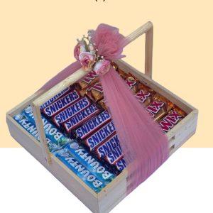 Symmetrically organized wooden chocolate tray filled with rows of Bounty, Snickers, and Twix bars, decorated with a pink net bow and roses